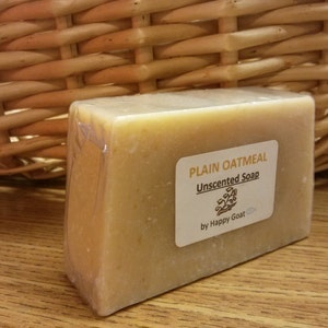 May include: A light beige, rectangular bar of unscented plain oatmeal soap, wrapped in clear plastic. The label reads "Plain Oatmeal Unscented Soap" with a logo and "by Happy Goat". The soap is displayed near a wicker basket.