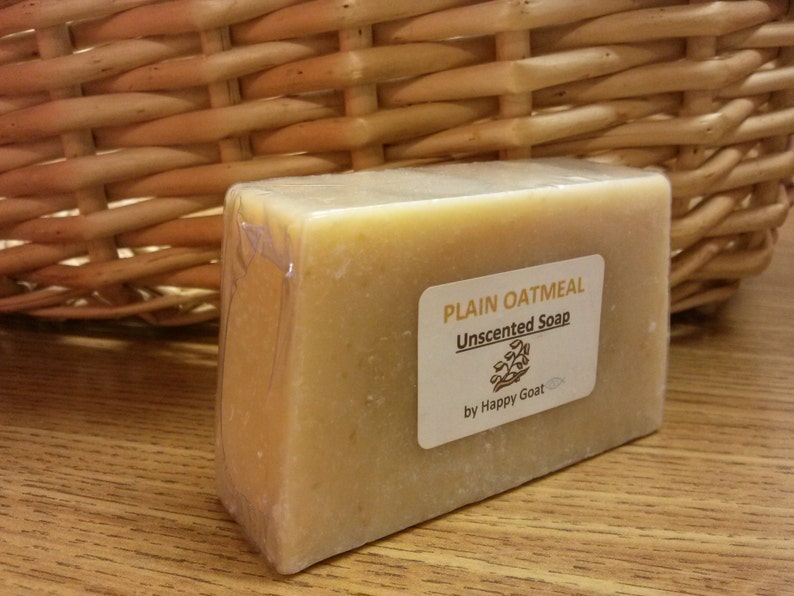 May include: A light beige, rectangular bar of unscented plain oatmeal soap, wrapped in clear plastic. The label reads "Plain Oatmeal Unscented Soap" with a logo and "by Happy Goat". The soap is displayed near a wicker basket.
