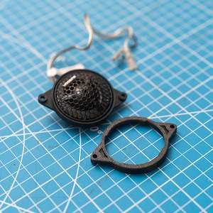 May include: A black speaker component with a honeycomb grill and a separate black mounting ring. The speaker has a wire attached. The items are on a blue grid surface.