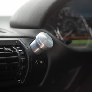 Könnte beinhalten: Ein zylindrischer Scheinwerferschalterknopf aus silbernem Metall, der an einem schwarzen Armaturenbrett neben einer horizontalen Lüftungsdüse angebracht ist. Der Knopf hat eine glatte, gebürstete Oberfläche über einem weißen Lichtsymbol.