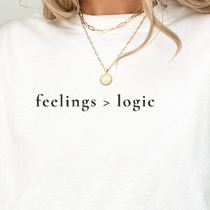May include: White sweatshirt with the text "feelings > logic" in a black font. The sweatshirt is a crew neck style and is being worn by a person. The person is wearing a gold necklace. The background is a neutral color.