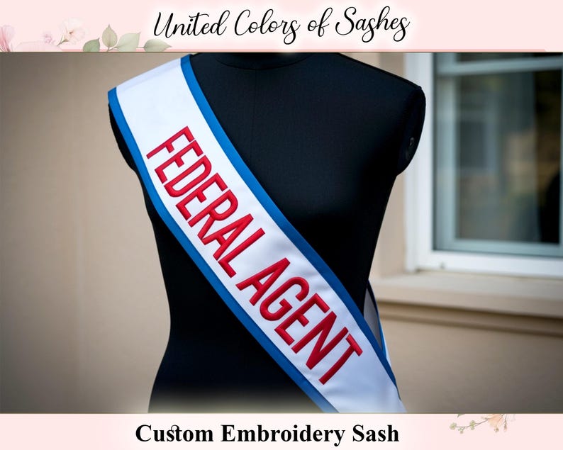 Op de afbeelding: Een witte sjerp met de woorden "FEDERAL AGENT" geborduurd in rood, omlijnd in blauw. De sjerp is over een zwarte mannequin gedrapeerd. De tekst "United Colors of Sashes" en "Custom Embroidery Sash" zijn ook zichtbaar.