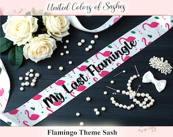 Op de afbeelding: Een witte satijnen sjerp met roze flamingo-illustraties en de tekst "My Last Flamingle" in het zwart. De sjerp is versierd met parels, een witte strik en een bloemenbroche. De woorden "United Colors of Sashes" en "Flamingo Theme Sash" zijn ook zichtbaar.