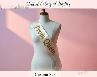 May include: A white satin sash draped over a mannequin, with the words "Prom Queen" in gold glitter lettering. The sash is a custom design, as indicated by the text "Custom Sash" at the bottom. The brand name "United Colors of Sashes" is at the top.