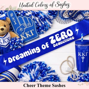 May include: A selection of cheerleading accessories in blue and white. Features a blue sash with the text "Dreaming of ZERO Deductions", a teddy bear, a foam hand, a tumbler, and pom-poms. The words "United Colors of Sashes" and "Cheer Theme Sashes" are also visible.
