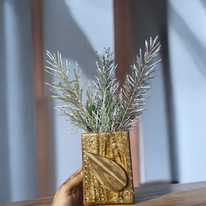 Könnte beinhalten: Eine rechteckige Holzvase mit Blattmuster und strukturierter Oberfläche. Die Vase enthält silberfarbene Zierzweige. Die Vase ist braun und steht auf einer Holzoberfläche.