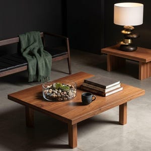 May include: A contemporary living room setting with a square wooden coffee table. It holds a glass bowl filled with stones and moss, books, and a black mug. A lamp with a stone base and a green throw are also present.