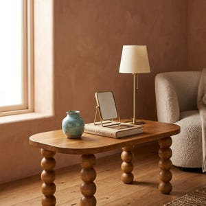May include: A wooden coffee table with a light brown finish, featuring a unique design with rounded legs. The table is adorned with a small lamp, a decorative vase, a mirror, and a book titled "Modernism in Design."