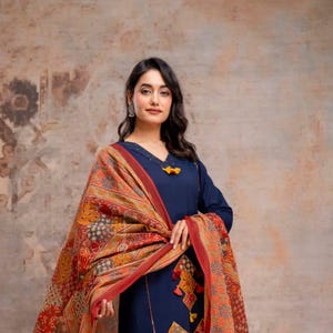 Puede incluir: Un conjunto de salwar kameez azul marino con un chal estampado. La túnica de manga larga presenta detalles decorativos y se combina con pantalones de pernera ancha. El chal tiene un borde rojo y un diseño geométrico colorido.