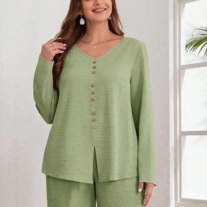 Puede incluir: Conjunto de dos piezas verde salvia. La parte superior tiene cuello en V, mangas largas y botones. Los pantalones a juego son de pierna ancha. El conjunto está hecho de tela texturizada.