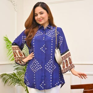 May include: A long-sleeved blue button-down shirt featuring a white geometric diamond pattern, a collar, and wide cuffs with a brown and cream paisley print. The shirt is paired with white trousers and a small cream-coloured shoulder bag with a gold chain.