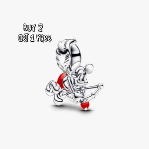 Pode incluir: Um charm de prata com um personagem de desenho animado como Cupido, segurando um arco e flecha com uma ponta de flecha em forma de coração vermelho. O personagem tem um detalhe em forma de coração vermelho nas costas. O texto "BUY 2 GET 1 FREE" está acima do charm.