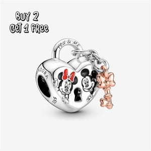 Pode incluir: Um berloque prateado em forma de coração com ilustrações de Mickey e Minnie Mouse. O berloque tem uma chave com um laço preso. O texto "YOU & ME" está na parte superior do berloque. O texto "BUY 2 GET 1 FREE" está no canto superior esquerdo.