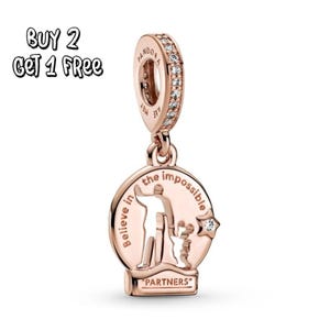 Pode incluir: Berloque em ouro rosa com um pingente redondo com a silhueta de uma pessoa e uma criança, com as palavras "Believe in the impossible" e "PARTNERS". O berloque tem uma faixa brilhante e a palavra "PANDORA".