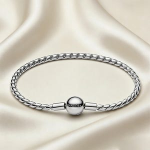 Puede incluir: Una pulsera plateada con un diseño de cadena trenzada. La pulsera presenta un cierre redondo con el nombre de la marca "Pandora" grabado. La pulsera se muestra sobre una superficie de satén blanco.