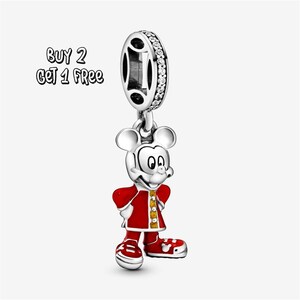 Pode incluir: Um berloque de prata com Mickey Mouse em um casaco e sapatos vermelhos com detalhes amarelos. O berloque tem um topo decorativo com pedras transparentes. O texto "BUY 2 GET 1 FREE" está no canto superior esquerdo.