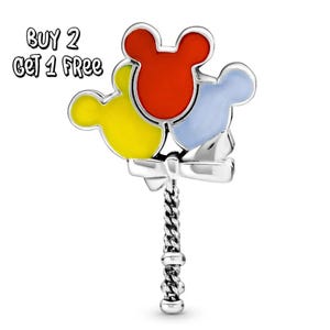 Pode incluir: Um berloque de prata com três balões em forma de cabeça do Mickey Mouse em vermelho, amarelo e azul. Os balões são conectados com um laço de prata e uma alça de prata torcida. O texto "BUY 2 GET 1 FREE" está no canto superior esquerdo.