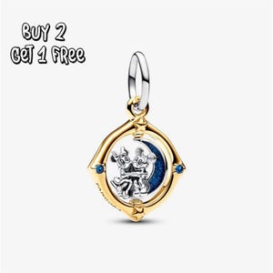 Pode incluir: Um charm dourado com um design de Mickey e Minnie Mouse em prata, abraçando-se em frente a uma lua crescente azul. O charm está definido em uma moldura quadrada com detalhes azuis e de estrelas. O texto "BUY 2 GET 1 FREE" está no canto superior esquerdo.