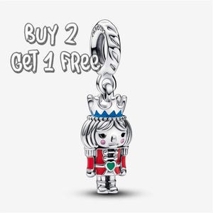 Puede incluir: Un charm de plata con un diseño de cascanueces. El charm presenta un uniforme rojo y plateado, una corona azul y un corazón verde. El texto "BUY 2 GET 1 FREE" está en la esquina superior izquierda.