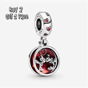 Pode incluir: Um berloque de prata com um design vermelho e branco. O berloque apresenta um pingente redondo com fundo vermelho e um coração, com duas figuras. A parte superior do berloque tem detalhes de coração vermelho. O texto "BUY 2 GET 1 FREE" está no canto superior esquerdo.
