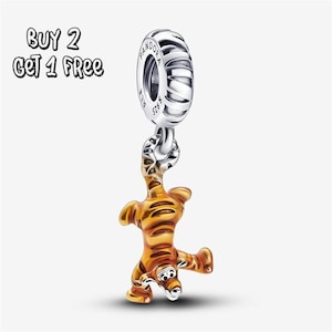 May include: A silver charm with a striped barrel design and a dangling orange and brown tiger figure. The charm is connected by a small chain. The text "BUY 2 GET 1 FREE" is in the upper left corner.