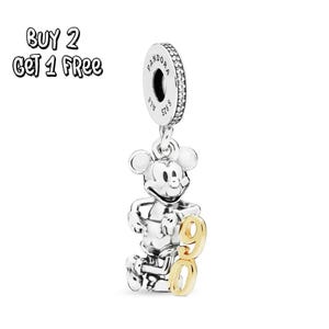 Pode incluir: Um berloque de prata com Mickey Mouse com um "90" dourado anexado. O berloque tem um topo circular com o nome da marca e "S925" gravado. O texto "BUY 2 GET 1 FREE" está no canto superior esquerdo.