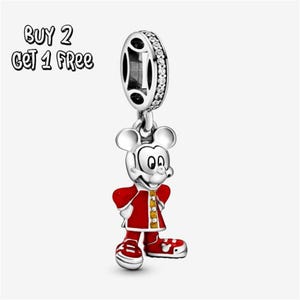 Pode incluir: Um berloque de prata com o Mickey Mouse em um casaco e sapatos vermelhos com detalhes amarelos. O berloque tem um design pendurado e um topo circular com pedras transparentes. O texto "BUY 2 GET 1 FREE" está no canto superior esquerdo.