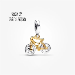 Puede incluir: Un charm de bicicleta dorado con ruedas plateadas y un lazo plateado para sujetarlo a una pulsera o collar. El texto "BUY 2 GET 1 FREE" está en la esquina superior izquierda.