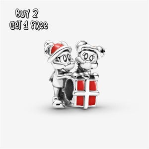 Pode incluir: Um berloque de prata com dois personagens de desenho animado, um com um chapéu vermelho e outro com um laço vermelho. Eles estão ao lado de uma caixa de presente vermelha. O texto "BUY 2 GET 1 FREE" está no canto superior esquerdo.