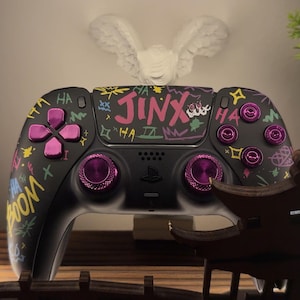 Könnte beinhalten: Schwarzer PlayStation 5-Controller mit individuellen Graffiti-Kunstwerken. Der Controller hat rosa Knöpfe und Akzente mit dem Wort "JINX" und anderen farbenfrohen Designs. Ein dekoratives Stück befindet sich im Vordergrund.