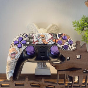 Może przedstawiać: Czarny kontroler PlayStation z fioletowymi przyciskami i niestandardowym wzorem z postaciami z kreskówek. Kontroler spoczywa na drewnianym elemencie dekoracyjnym z motywem mostu, oświetlonym okrągłym światłem.