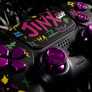 Op de afbeelding: Zwarte gamecontroller met paarse knoppen en accenten. De controller heeft het woord "JINX" in roze en witte graffiti-letters. Er zijn ook extra kleurrijke graffiti-ontwerpen aanwezig.