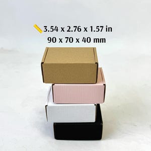 May include: A stack of four rectangular cardboard boxes in brown, light pink, white, and black. The text above the boxes indicates dimensions of 3.54 x 2.76 x 1.57 inches. Each box features a hinged lid design with a flat top and vertical sides.