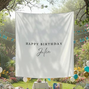 May include: A white banner with the text "HAPPY BIRTHDAY Julia" hangs in a garden setting. The scene includes balloons, chairs, a table with a cake, and gift boxes, suggesting a birthday celebration. The banner is the focal point.
