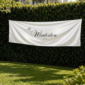 Könnte beinhalten: Ein weißes Satinbanner mit dem Text "the Winterton" in eleganter schwarzer Schrift, zusammen mit dem Datum 28.09.2020. Das Banner wird im Freien vor einem Hintergrund aus grünem Laub und Gras präsentiert, was auf eine festliche Veranstaltung hindeutet.