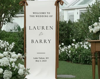Könnte beinhalten: Ein weißes Hochzeits-Willkommensschild mit braunen Holzstützen und Bändern. Auf dem Schild steht "WELCOME TO THE WEDDING OF LAUREN & BARRY" und enthält Datum und Ort: Lake Tahoe, NV, 3. Mai 2025.