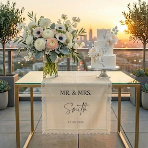 May include: A three-tiered white wedding cake with floral decorations sits on a silver stand. A table runner with the text "MR. & MRS. Smith" and the date 17.04.2026 is displayed. A bouquet of flowers and greenery is in a glass vase.
