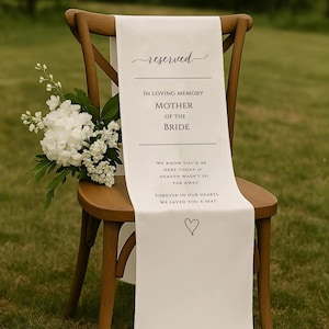 May include: A wooden chair with a white fabric runner featuring the words "reserved" and "In Loving Memory Mother of the Bride." A bouquet of white flowers is placed on the chair. A heart symbol is at the bottom of the runner.
