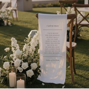 Puede incluir: Una pancarta de tela blanca con el texto "I will be there" y un poema, sobre una silla de madera. La escena incluye flores blancas, velas y otras sillas, en un césped, ideal para una boda.