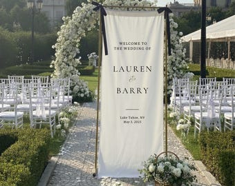 Könnte beinhalten: Ein weißes Hochzeitsbanner mit goldenem Rahmen und schwarzen Akzenten. Auf dem Banner steht "WELCOME TO THE WEDDING OF LAUREN & BARRY" und enthält Ort und Datum: Lake Tahoe, NV, 3. Mai 2025. Ein Blumenbogen und Stühle sind im Hintergrund.