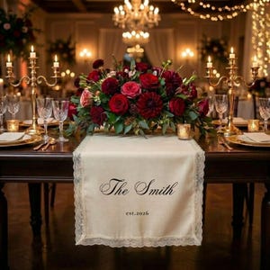 Könnte beinhalten: Eine festliche Tischdekoration mit einem cremefarbenen Tischläufer mit der Aufschrift "The Smith" und "est.2026". Der Läufer ist mit Spitze verziert. Ein Blumengesteck aus roten Rosen und Dahlien befindet sich darauf. Goldene Kerzenhalter und Gedecke vervollständigen die elegante Einrichtung.