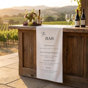 May include: A rustic wooden bar with a white linen runner displaying "The Bar" and cocktail recipes. Bottles of wine, glasses, and a floral arrangement are arranged on the bar. The background features a vineyard.