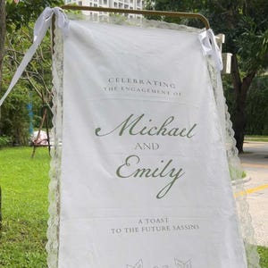 Puede incluir: Una pancarta de compromiso blanca con ribete de encaje y un marco dorado. La pancarta dice "Celebrating the Engagement of Michael and Emily" en una elegante escritura, con una ilustración de paloma. Cintas blancas atadas en la parte superior.