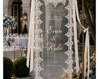 May include: A vertical rectangular wedding welcome sign made of sheer fabric with intricate white lace borders. The sign features the text "WELCOME TO THE WEDDING OF Evan & Rylie La Savole February 1, 2026" and is suspended by cream-colored ribbons.