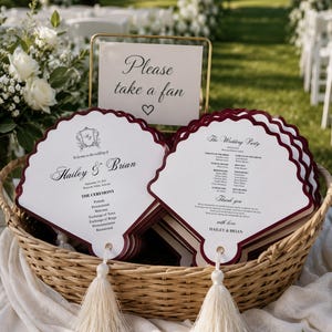 May include: A wicker basket filled with scalloped-edge wedding program fans featuring deep red borders, white cardstock, and cream-colored tassels. A gold metal sign holder displays a card reading Please take a fan with a heart icon.
