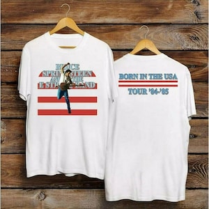 Puede incluir: Camiseta blanca con un diseño gráfico de Bruce Springsteen and the E Street Band, con detalles en rojo y azul. El frente muestra a Springsteen tocando la guitarra. La parte trasera de la camiseta dice "Born in the USA" y "Tour '84-'85".