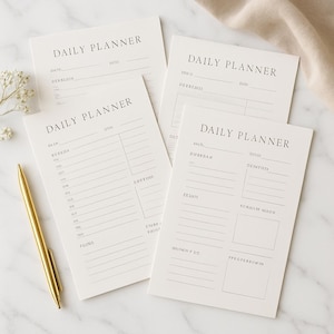 Puede incluir: Cuatro blocs de notas de planificación diaria de color blanquecino con las palabras "DAILY PLANNER" impresas en la parte superior. Un bolígrafo dorado y pequeñas flores blancas están a la izquierda. Los blocs de notas tienen líneas para notas y planificación.