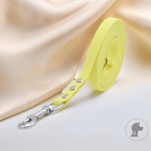 Op de afbeelding: Een opgerolde, heldergele hondenriem met een getextureerd oppervlak en zilverkleurige hardware. De riem is gemaakt van een duurzaam materiaal en heeft een metalen sluiting voor eenvoudige bevestiging aan de halsband van de hond. De riem ligt op een witte ondergrond.
