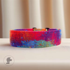 Peut inclure: Un collier pour chien coloré avec un motif abstrait. Le collier présente un mélange de teintes rouges, bleues et jaunes, créant un effet tie-dye ou aquarelle. Il est équipé d'une boucle noire et d'un anneau en D pour l'attache de la laisse.
