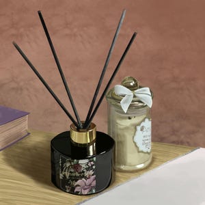 Puede incluir: Un difusor de aromaterapia negro con varillas y un collar dorado, junto a una vela en tarro de cristal con un lazo blanco. El difusor tiene una etiqueta floral con la palabra "aromaterapia". La vela tiene una etiqueta con texto.